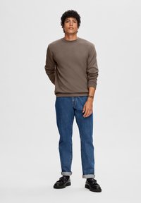 Bruine sweatshirt met ronde hals, gecombineerd met blauwe spijkerbroek. De outfit is compleet met zwarte schoenen en opgerolde manchetten op de broek.