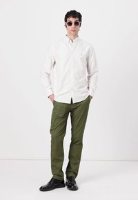 Camisa de manga larga a rayas blancas, pantalones verde oliva, zapatos negros, con gafas de sol, de pie contra un fondo blanco liso.
