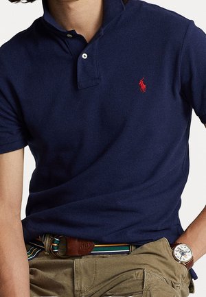 Uomo che indossa una polo blu navy con logo ricamato rosso, cintura a righe, pantaloni kaki e orologio da polso con quadrante bianco e cinturino in pelle marrone.
