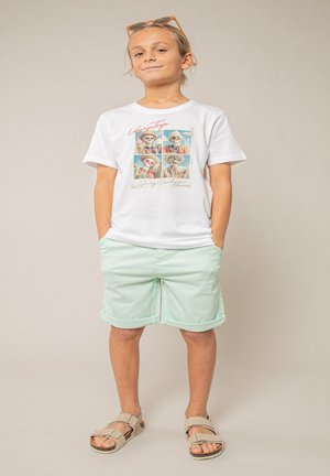 Enfant debout les mains dans les poches, portant un t-shirt blanc avec un motif de squelettes, un short vert menthe, des sandales beiges et des lunettes de soleil orange sur la tête.