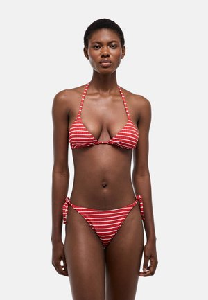 Femme portant un haut de bikini rayé rouge et blanc à col halter et un bas de bikini assorti avec liens latéraux, debout devant un fond clair uni.