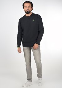Dark gray sweatshirt with round neckline, ribbed cuffs, and hem. Features small green logo on the chest. Worn with gray jeans and white sneakers.