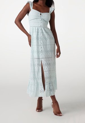 Robe longue bleu clair en tissu ajouré, décolleté carré et détails de boutons. Dispose d'une fente frontale et d'un ourlet à volants. Portée avec des talons marron.