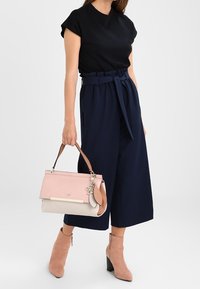 Sac à main rose clair et crème avec une bandoulière marron, doté d'un accent doré et d'un charm. Porté avec un haut noir et un pantalon palazzo bleu marine.