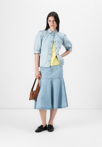 A woman stands confidently in a light blue denim jacket and skirt, paired with a bright yellow top and black shoes, holding a brown bag.
