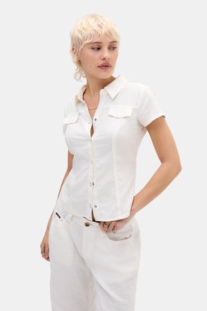 Moteris trumpais šviesiais plaukais, dėvinti prigludusią baltą trumpomis rankovėmis palaidinę su sagomis ir laisvas baltas kelnes, žiūrinti į šoną.