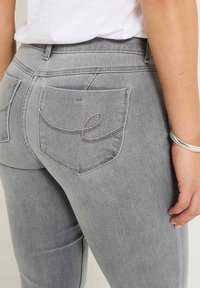 Vaqueros de mezclilla gris con corte slim. Presentan dos bolsillos traseros con costura curva y una cinturilla visible, mostrando un detalle sutil.