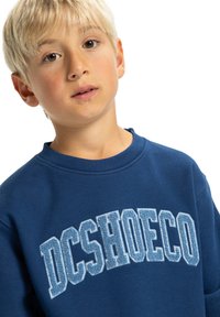 Modrá mikina vyrobená z mäkkej tkaniny, s výrazným bielym vyšívaným textom "DCSHOECO" s textúrovaným vzorom. Okrúhly výstrih a dlhé rukávy.