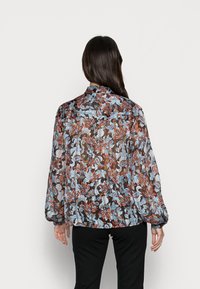Blusa em tecido transparente, com padrão floral, apresentando mangas longas e punhos elásticos em tons de azul, laranja e castanho sobre um fundo translúcido.