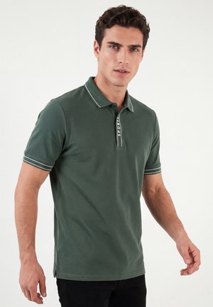 Młody mężczyzna w ciemnozielonej koszulce polo z białym wykończeniem i napisem "SPORT" na listwie z guzikami, stojący na jednolitym tle.