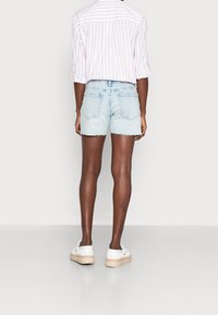 Ljusblå denimshorts med fransad nederkant, tillsammans med en randig rosa och vit skjorta, sedd bakifrån. Vita sneakers fulländar outfiten.