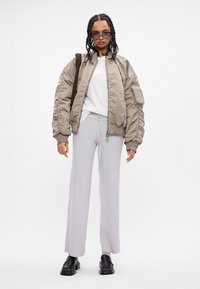 Casaco bomber bege claro com tecido texturizado e bolsos laterais, combinado com calças largas cinza claro e sapatos pretos. Design minimalista, sem padrões.