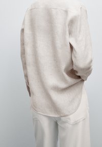 Jasno-beżowa lniana koszula o lekko teksturowanym wykończeniu, z zaokrąglonym dołem i długimi rękawami, widok z tyłu.