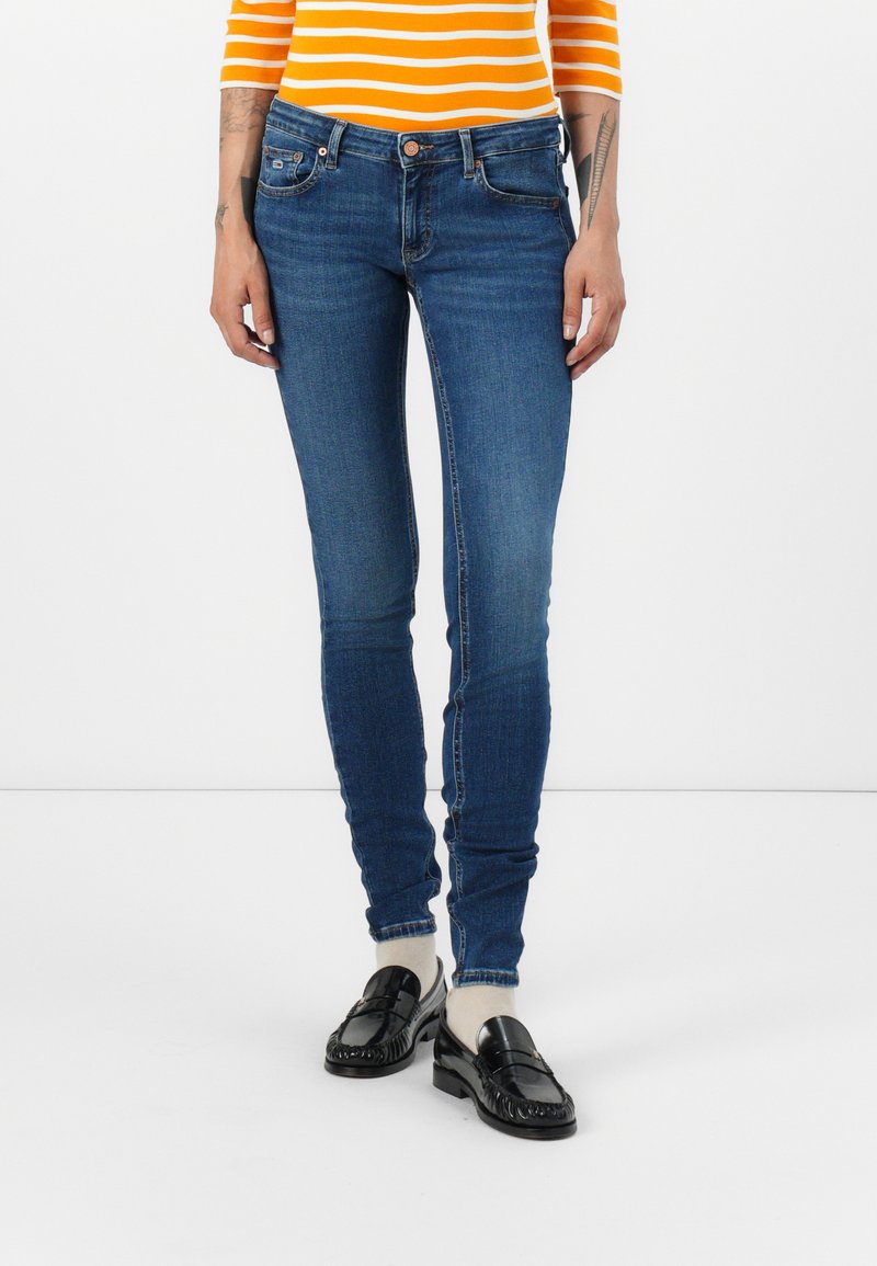 Osoba ubrana w niebieskie obcisłe dżinsy, czarne mokasyny, białe skarpetki i pomarańczowo-białą pasiastą bluzkę, stojąca na tle białym.