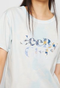 Kvinna bär en ljusblå tie-dye T-shirt med blommönstrad text "See By Girl" över bröstet.