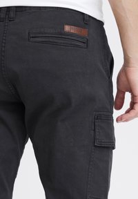 Primo piano di una persona che indossa pantaloni cargo neri con tasche laterali e posteriori e una etichetta di pelle sulla cintura.