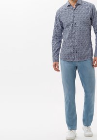 Hombre vistiendo una camisa de botones de manga larga azul con flores, jeans de mezclilla azul claro y zapatillas blancas, de pie contra un fondo blanco liso.