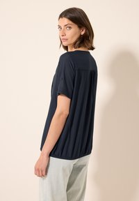 Maglietta blu navy a maniche corte, tessuto strutturato e design con schiena arricciata. Abbinata a pantaloni chiari per un look casual.