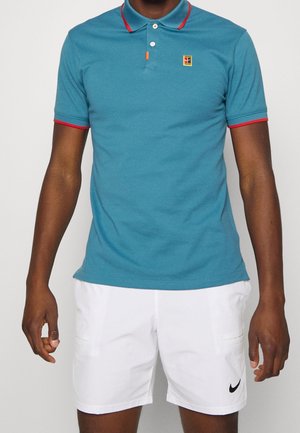 Homme portant un polo bleu sarcelle avec bordure rouge et un short blanc Nike, debout devant un fond uni.