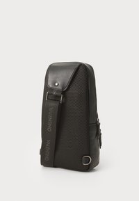 Borsa nera con un design a rete texturizzato, chiusura con zip, parte superiore piatta e una tracolla regolabile con logo. Accenti lisci e minuteria in metallo.