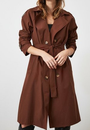 Vrouw die een lange bruine trenchcoat met riem draagt, met opgerolde mouwen en grote knopen, handen samengevouwen ter hoogte van de taille.