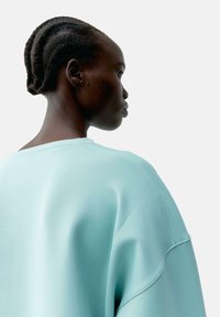 Felpa oversize azzurra chiaro con una superficie liscia, scollatura ampia e dettagli cuciti. Capelli del modello acconciati in trecce, mostrati di profilo.