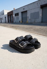Sandales compensées noires avec doubles brides pailletées et boucles argentées, posées sur un trottoir de ville près de bâtiments industriels.