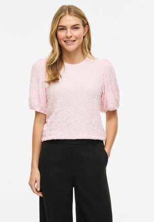 Smaidoša sieviete ar blondiem matiem valkā gaiši rozā teksturētu augšu ar pūkainām piedurknēm un melnas bikses, stāvot ar vienu roku kabatā.