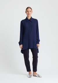 Navyblaues Langarmhemd mit spitzem Kragen, durchgehender Knopfleiste und asymmetrischem Saum, kombiniert mit dunklen Hosen und metallischen Schuhen.