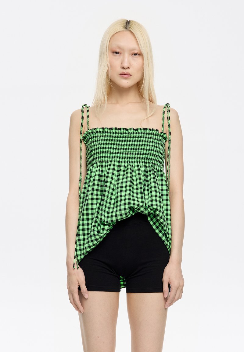 Zelena in črna gingham crop majica s smockanim bodicem, nastavljivimi zavezicami in lepršavim robom; nošena z črnimi kratkimi hlačami.