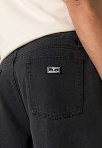 Black denim pants with a back pocket featuring a rectangular brand label. Fabric has a textured surface with subtle stitching details.