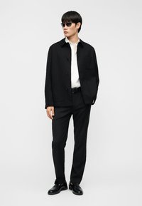 Νεαρός άνδρας που στέκεται, φοράει μαύρα γυαλιά ηλίου, μαύρο σακάκι και παντελόνι, λευκό πουκάμισο, μαύρη ζώνη και μαύρα παπούτσια επίσημου τύπου σε απλό φόντο.