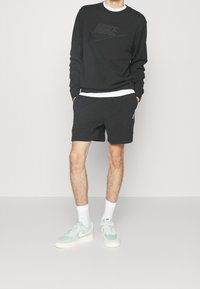 Man som bär en svart Nike-tröja och shorts, vita strumpor, och ljusgröna och vita Nike-sneakers, står med händerna i fickorna.