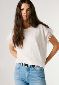 Młoda kobieta z długimi brązowymi włosami, ubrana w biały top z frędzlami na rękawach oraz jasnoniebieskie dżinsy z czarnym paskiem, na jednolitym tle.