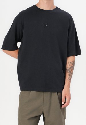 Persona que lleva una camiseta negra holgada con un pequeño logo en el pecho y pantalones verde oliva, mostrando un tatuaje en el antebrazo izquierdo.