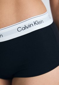 Sorte bomullstruser med en hvit elastisk midjebånd som har "Calvin Klein"-logoen i svart tekst, glatt tekstur og figursydd design.