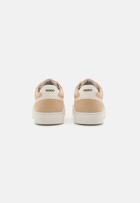 Baskets en daim beige avec semelles en caoutchouc blanc, présentant un talon arrière surélevé et un logo HUGO sur l'étiquette du talon. Design épuré et minimaliste.