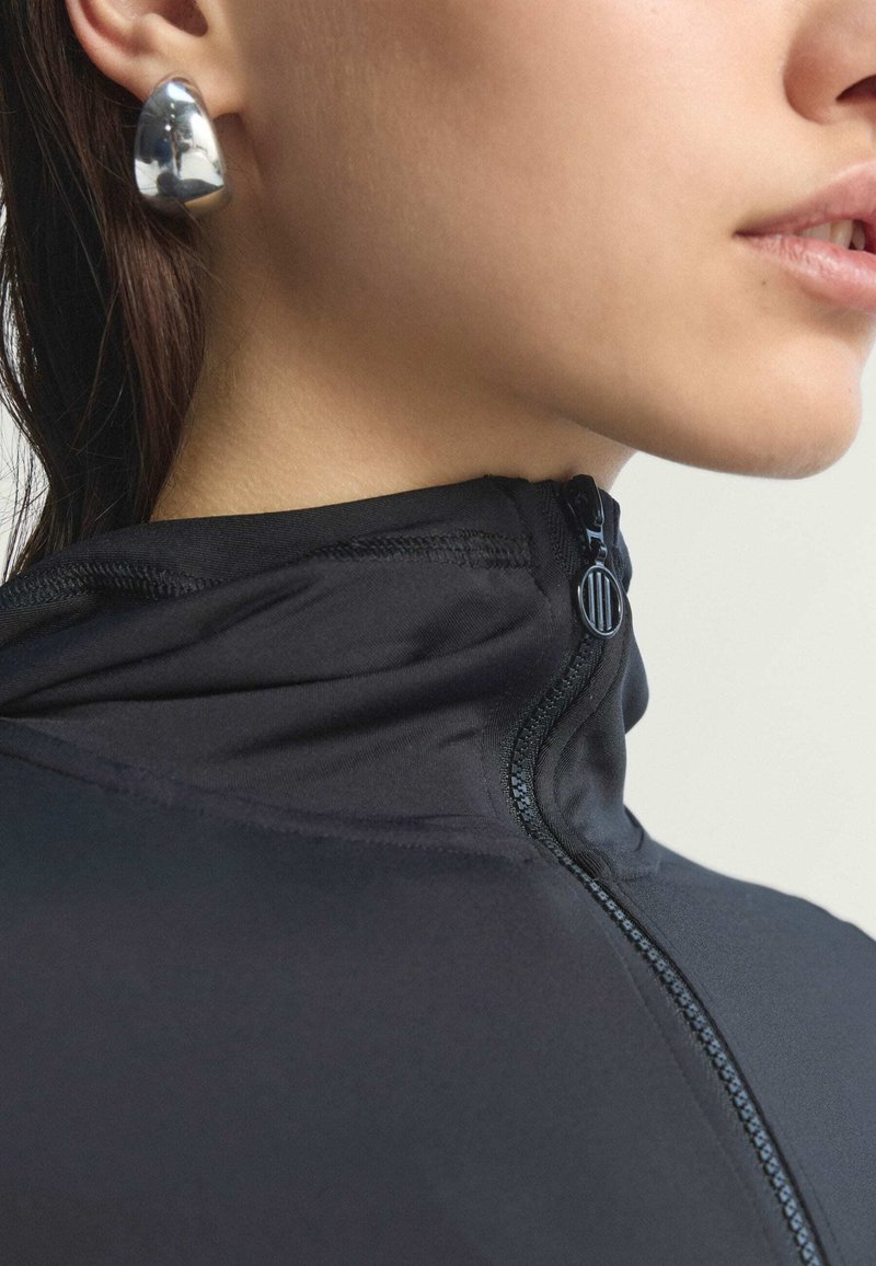 Giacca nera con zip e colletto alto, caratterizzata da una texture liscia. Orecchino scultoreo argentato e lucido che accentua l'orecchio.