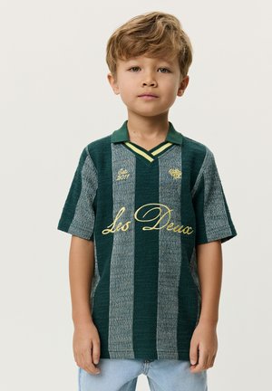 Jeune garçon debout, portant un polo à manches courtes rayé vert et gris avec l'inscription jaune « Les Deux » et un jean bleu clair.