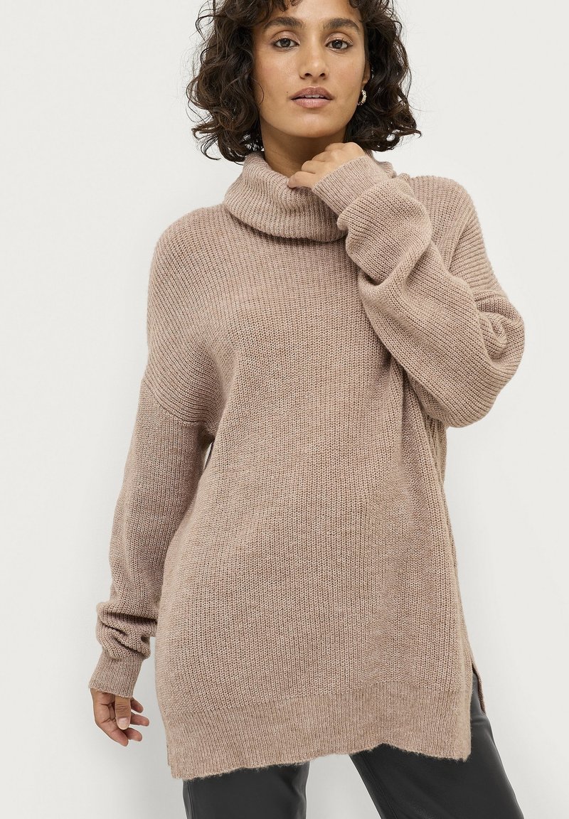Beige, oversized sweater med en hættekrave, lavet af et blødt, struktureret strikmateriale. Har drop-skulder og ribbede manchetter.