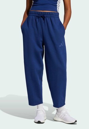 Personne portant un pantalon de jogging bleu marine à jambes larges, debout les mains dans les poches, associé à des chaussures de sport blanches et des chaussettes.