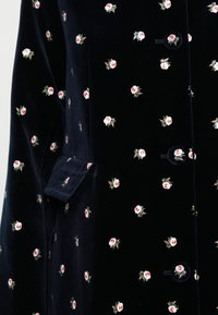 Dunkelveloursstoff mit kleinen rosa und weißen gestickten Rosenmustern, der Knöpfe und eine Klapptasche auf dem Kleidungsstück zeigt.