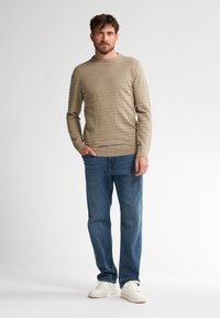 Beiger, strukturierter Pullover mit horizontalen Rippungen, Rundhalsausschnitt; kombiniert mit mittelblauen Jeans und weißen Sneakers. Minimalistisches Design und neutrale Farben.