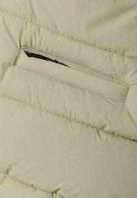 Helle grüne Daunenjacke aus einem Stoff mit sichtbaren Nähten und einer Reißverschlusstasche. Die Textur wirkt weich und leicht knittrig.