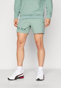 Pantalones cortos deportivos color verde menta con un logo de PUMA, combinados con zapatillas blancas que tienen acentos en rojo y negro y calcetines blancos texturizados.