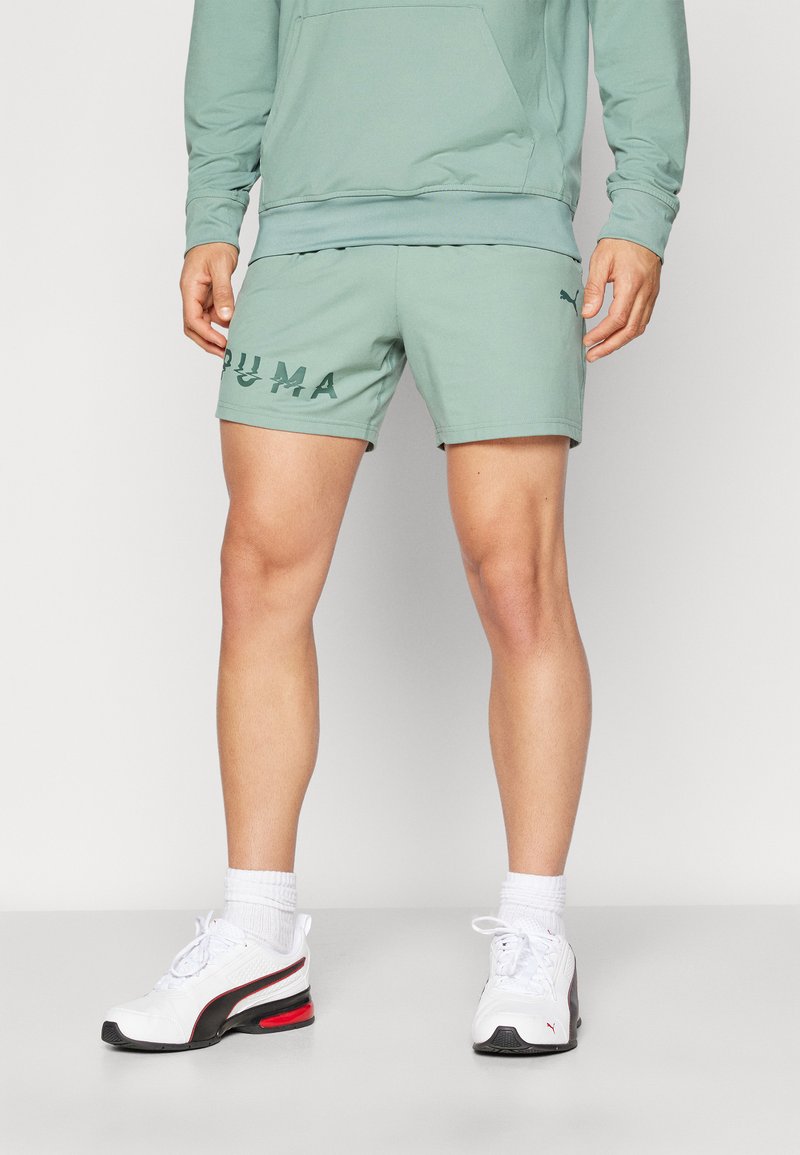 Pantalones cortos deportivos color verde menta con un logo de PUMA, combinados con zapatillas blancas que tienen acentos en rojo y negro y calcetines blancos texturizados.