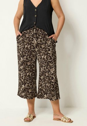 Kvinde iført sort ærmeløs top, brune leopardmønstrede vide knickers og guld flade sandaler, står med hænderne i lommerne.