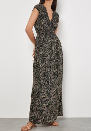 Woman wearing a black maxi dress with green and beige leaf print, cap sleeves, ruffled neckline, standing against a white wall.