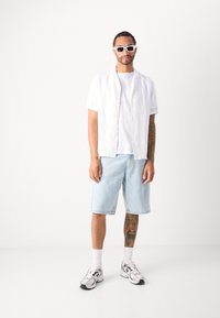 Hvit kortermet skjorte med knapper over en hvit t-skjorte, blå denimshorts, hvite sokker, sølvfargede joggesko og hvite rektangulære solbriller.
