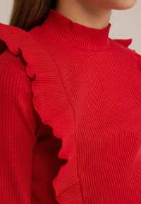 Červený rebrovaný turtleneck s volánovými akcentmi na pleciach. Textúrovaný, mierne lesklý materiál. Detailný pohľad na dizajn.